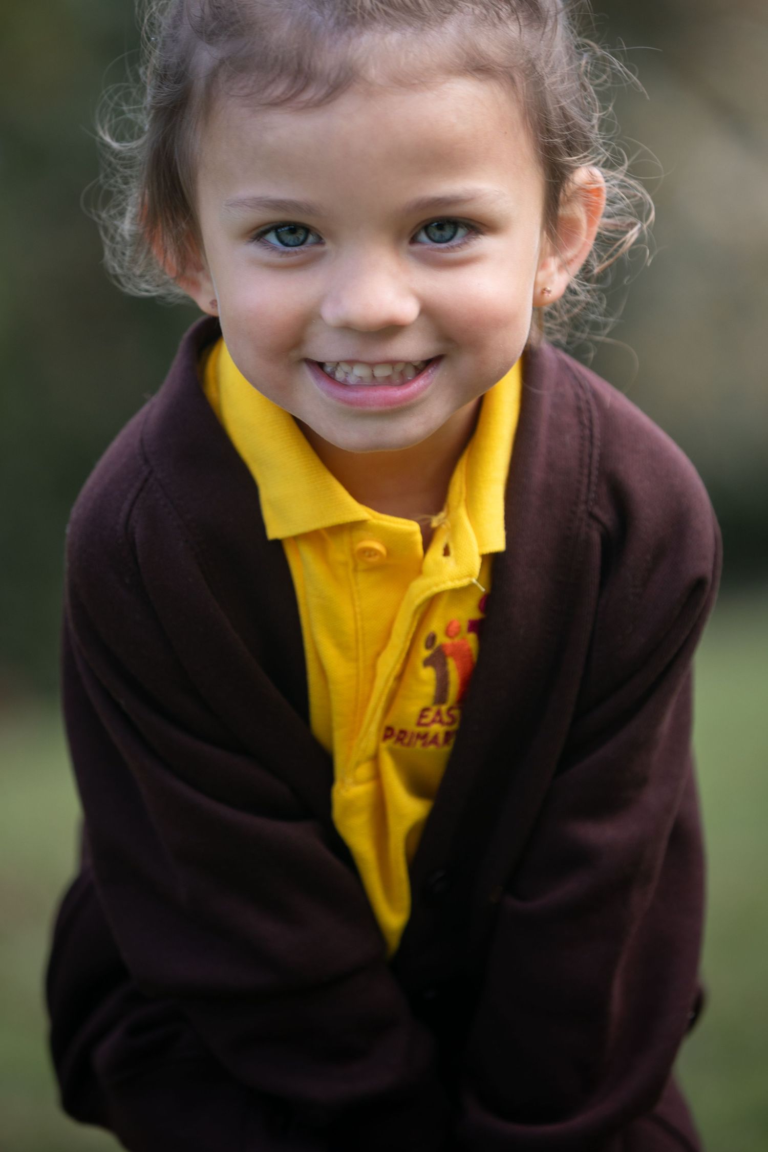 Smiling young girl in yellow school polo shirt and dark maroon fleece jacket posing outdoors.