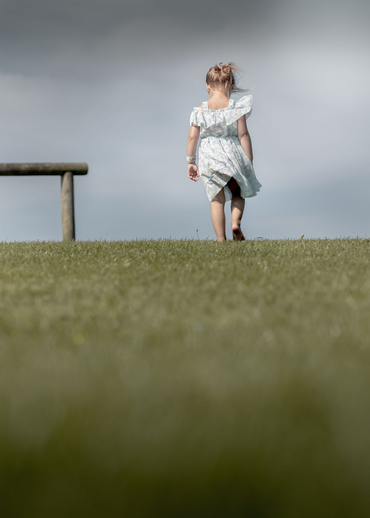 Young girl in white dress walking on green grass field with wooden bench and cloudy sky backdrop.