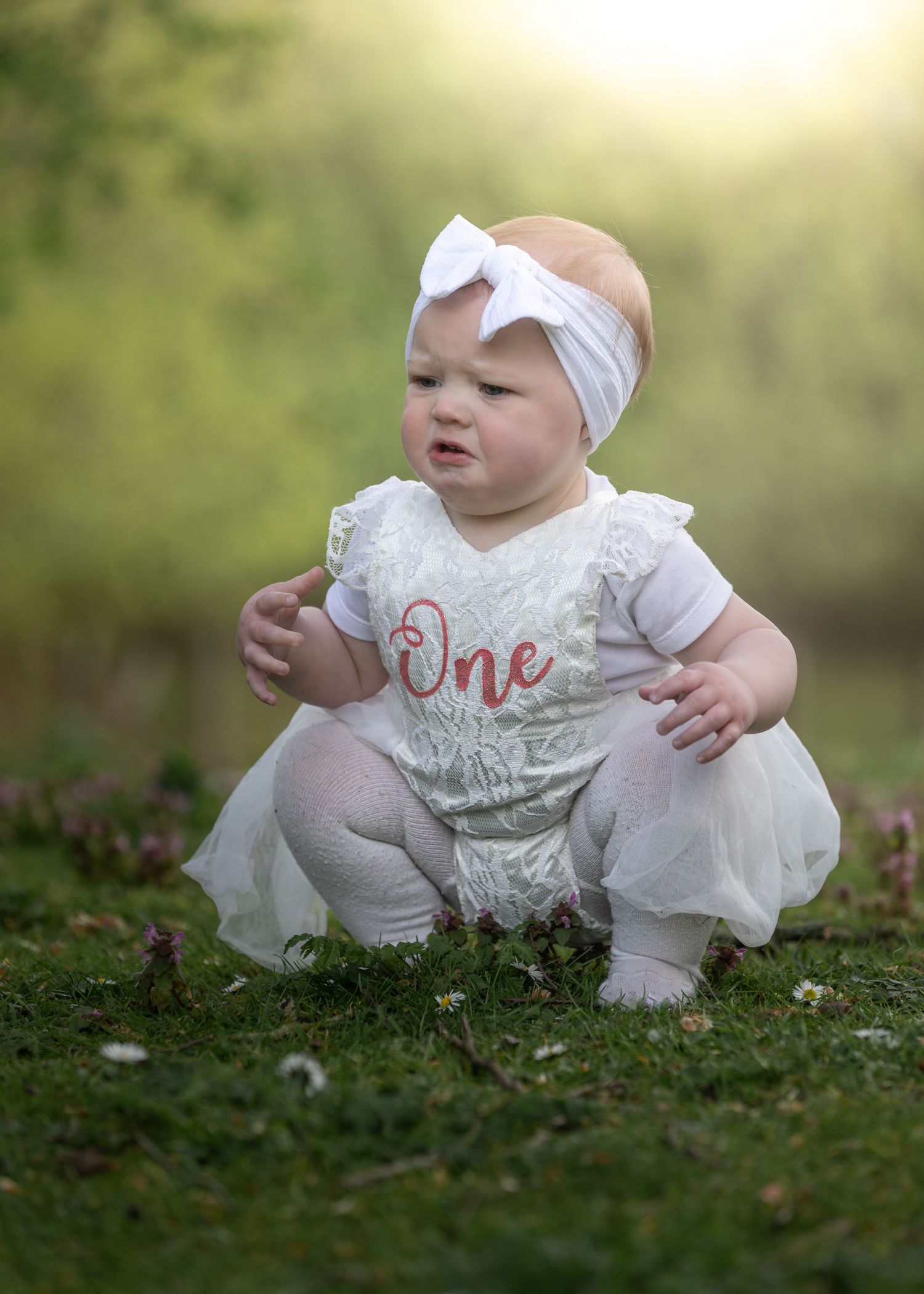 Baby girl in white lace One birthday romper and bow headband sitting on grass in soft golden light.

East Park Hull
