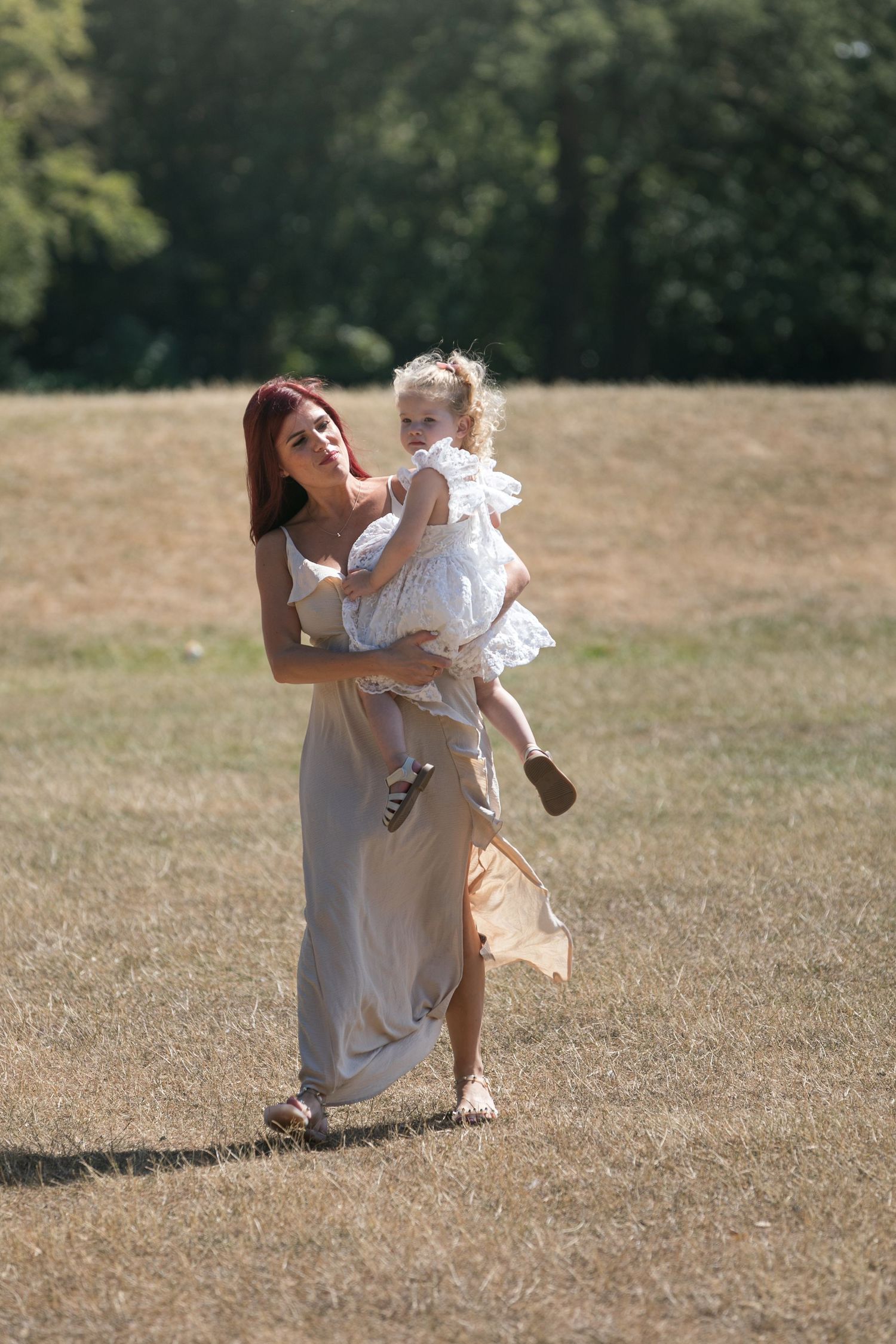 A mother in a beige dress holds her toddler daughter in a sunny park field with green trees in the background.