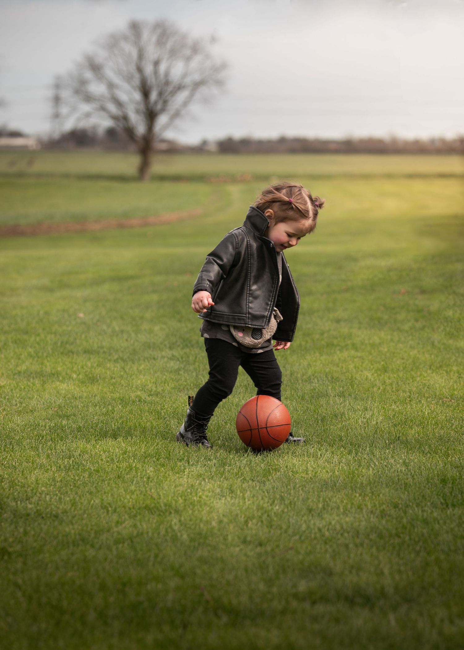 Young child in black leather jacket kicking a basketball on a green grass field.