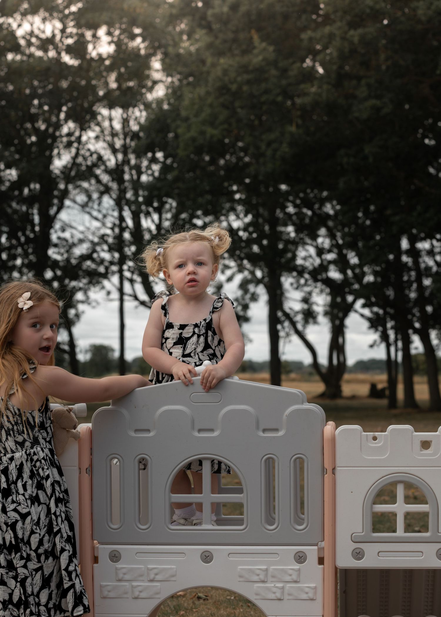 Two young girls playing on a plastic outdoor playhouse in a park surrounded by trees.