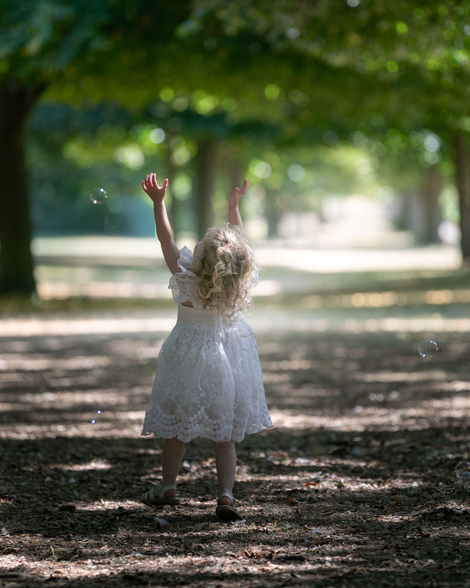 A young girl in a white lace dress reaches up joyfully along a tree-lined path in a sunlit park.