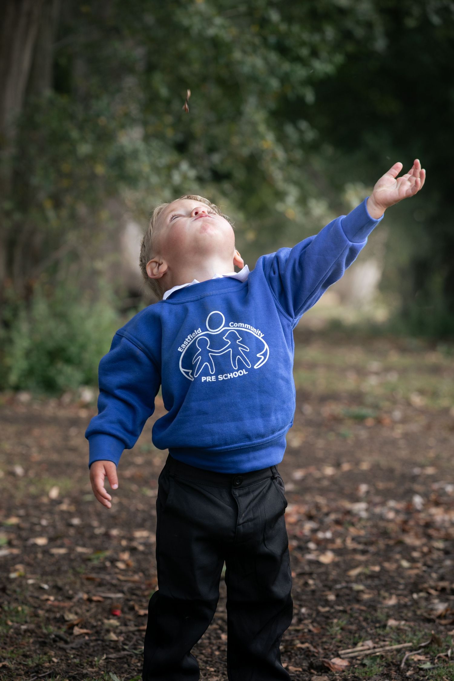 A young toddler in a blue preschool sweatshirt reaches up toward the sky outdoors among fallen leaves.