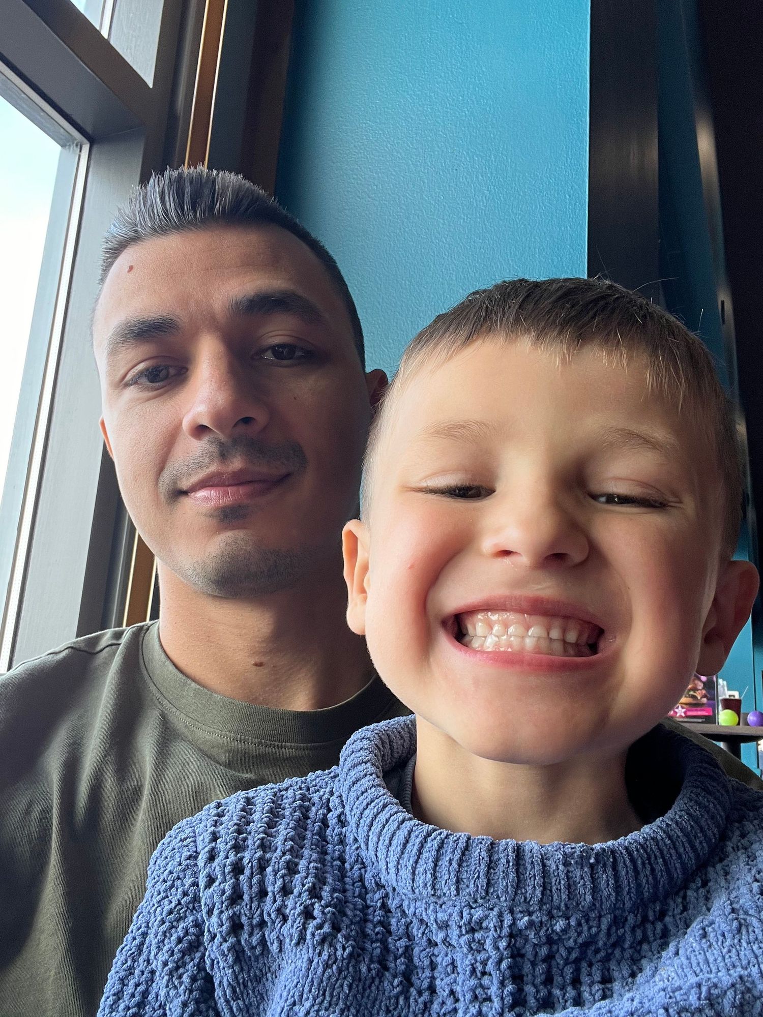 Father and young son smiling together for a selfie indoors near a window.