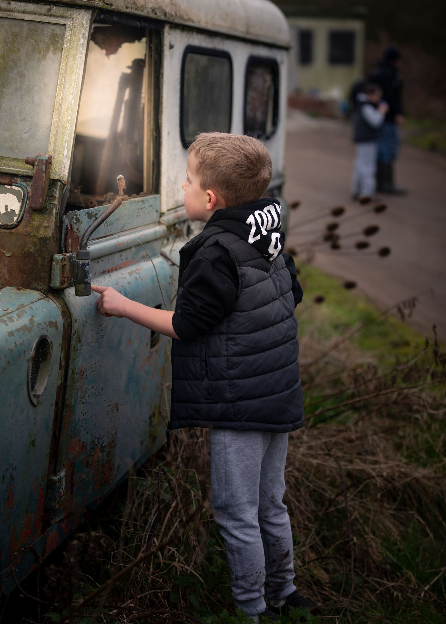 Young boy in black vest examining a rusty vintage truck outdoors, with blurred figures visible in the background.
