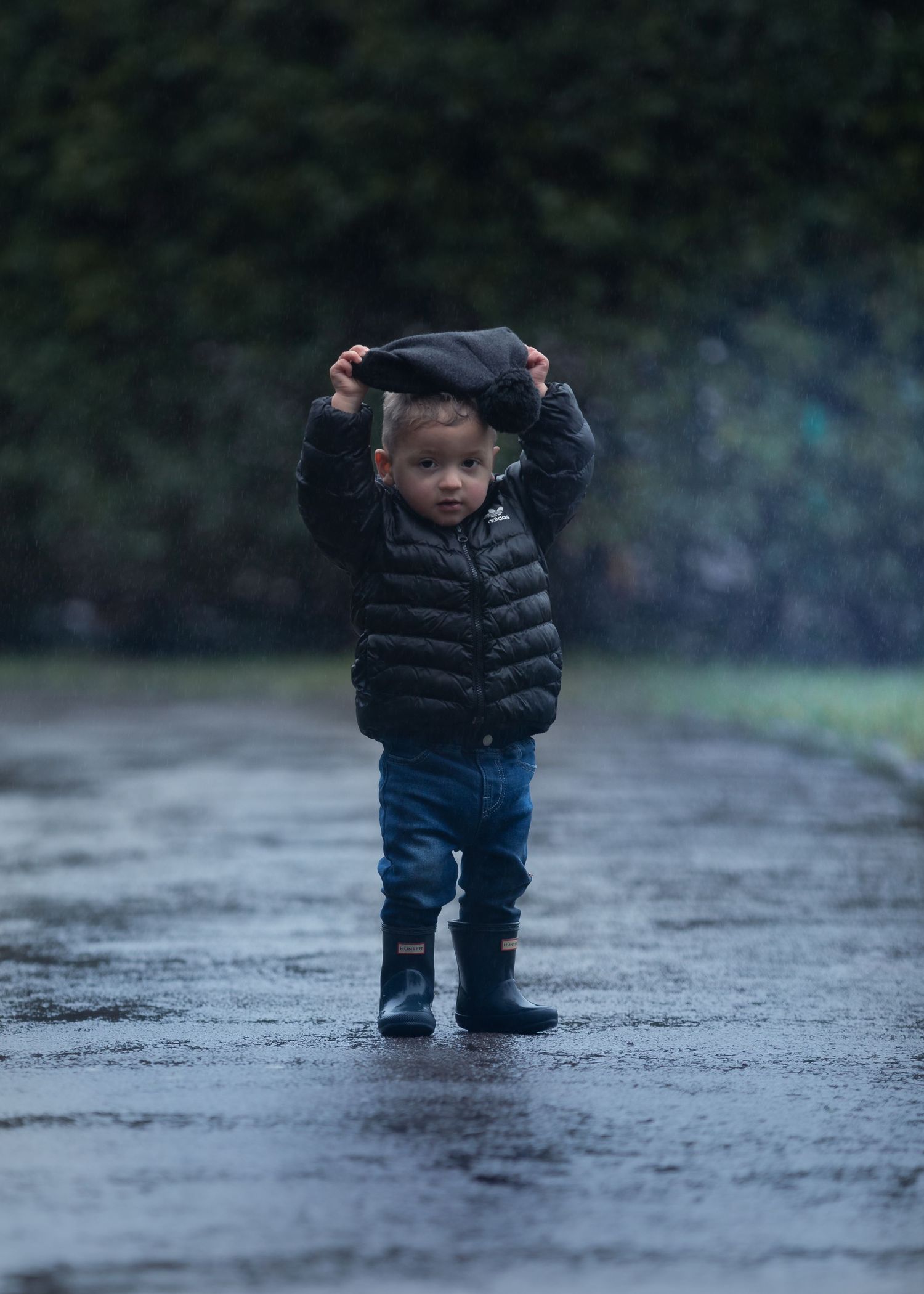Toddler boy in black puffer jacket pulling hood over head while standing in rain on wet pavement.

Sutton Village