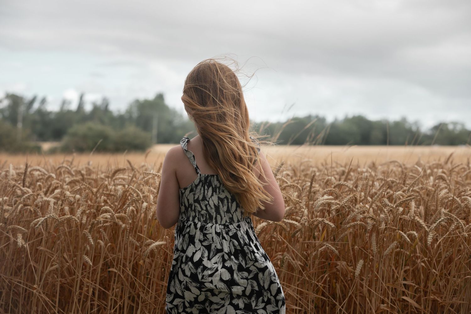 Woman in floral dress standing in golden wheat field with hair blowing in wind on overcast day.