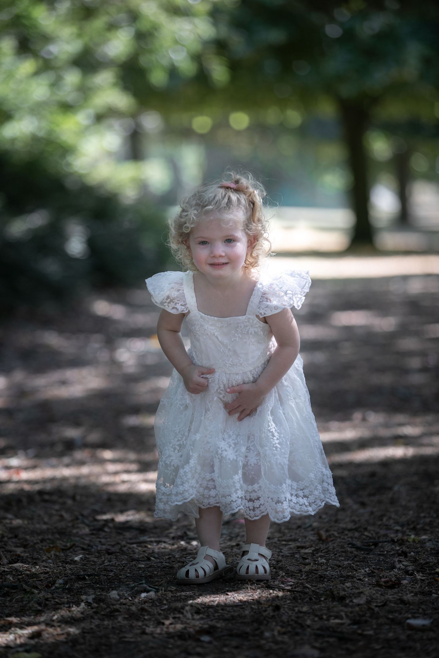 Smiling toddler girl in white lace dress with curly blonde hair walking on a shaded park path.