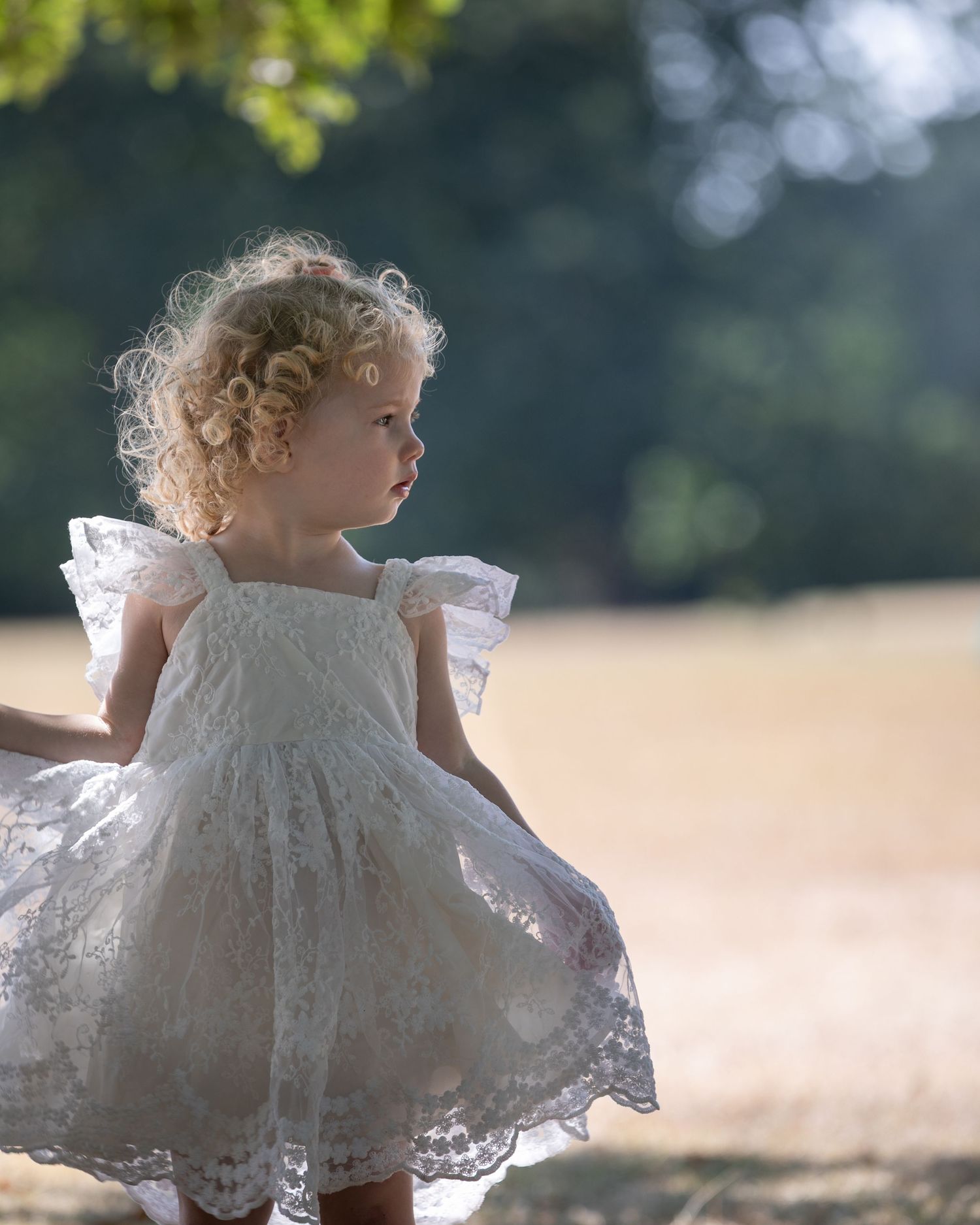 A toddler girl with curly blonde hair wearing a white lace dress twirls outdoors in a sunlit field.