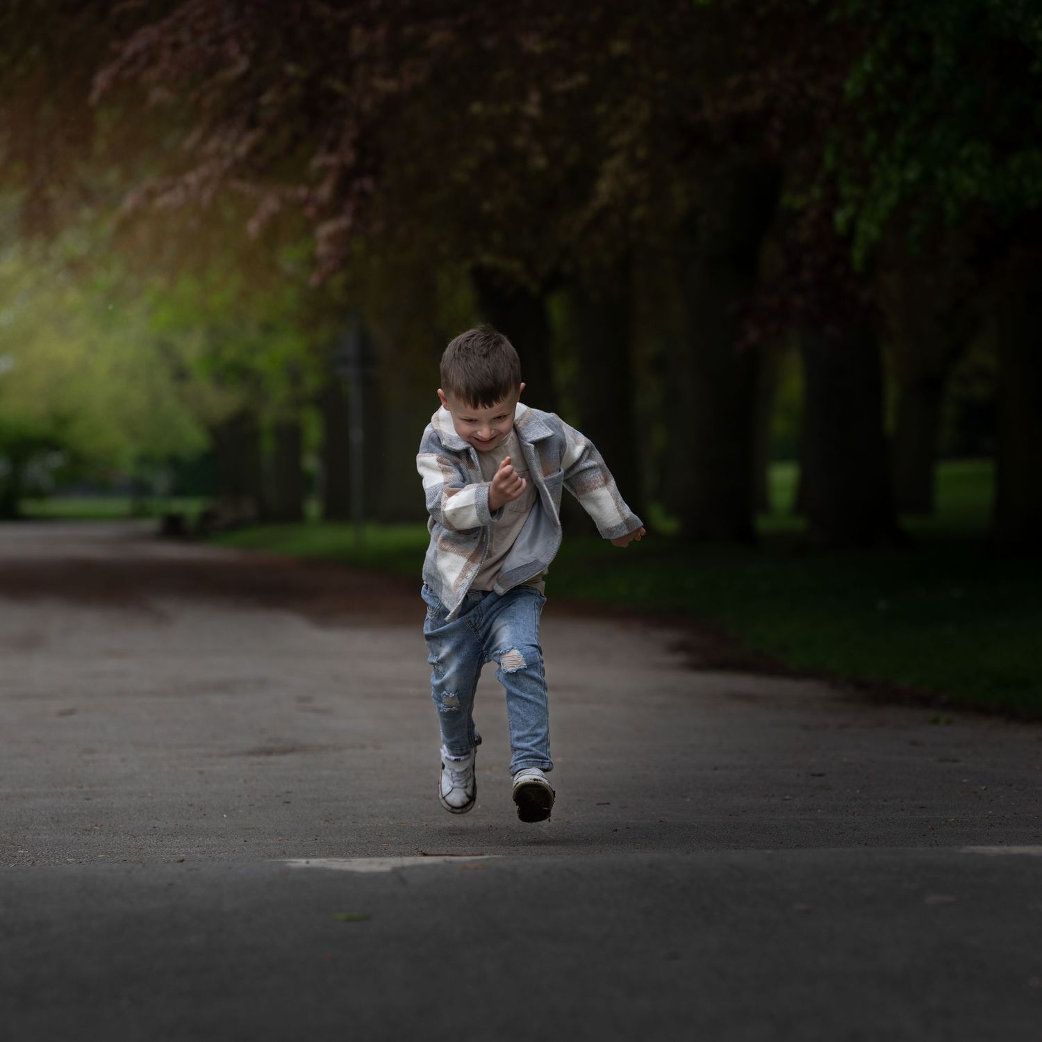 A young boy runs energetically down a tree-lined park path, wearing a plaid jacket and ripped jeans.
