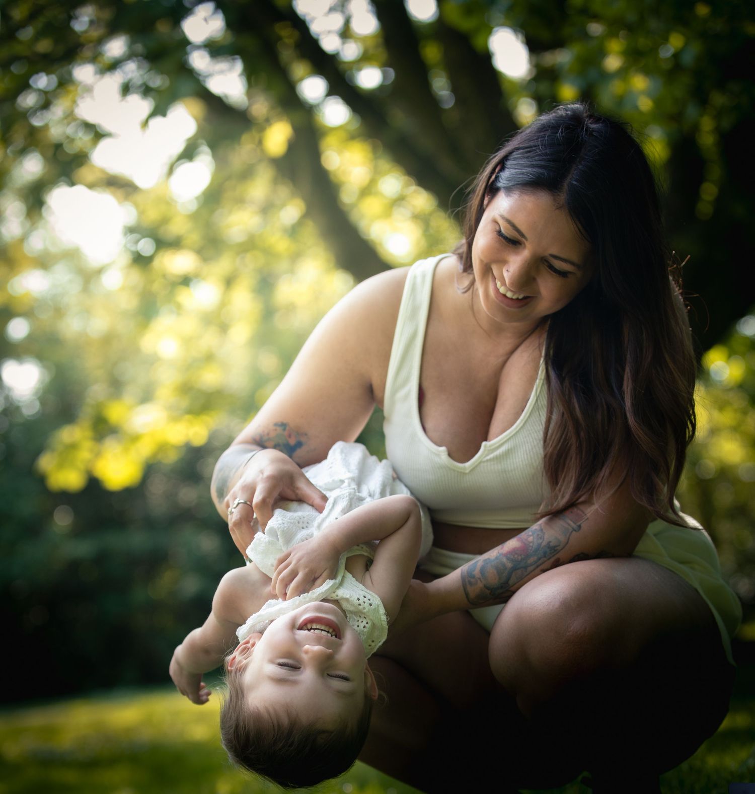 A smiling tattooed mother playfully holds her laughing baby upside down in a sunlit green park.