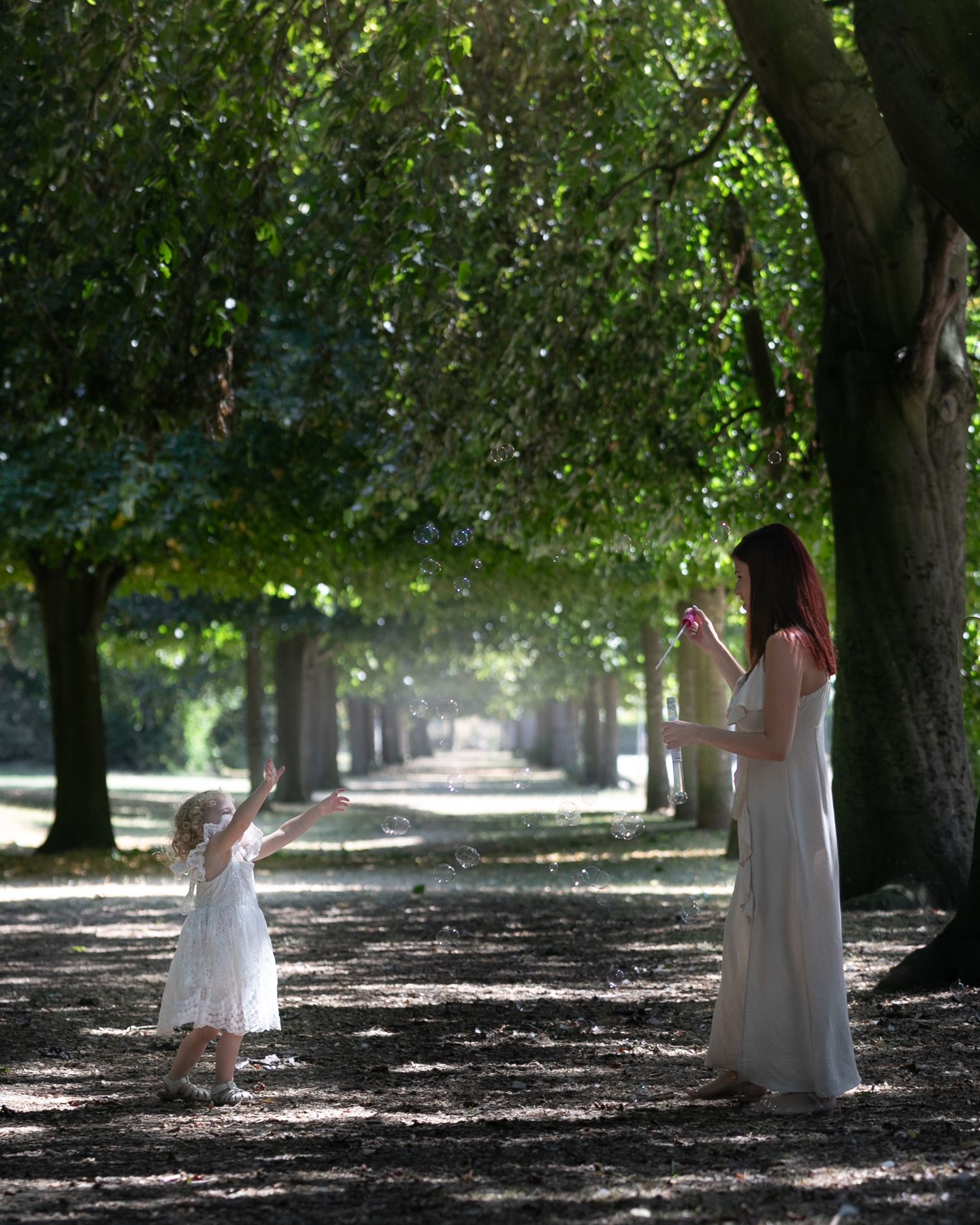 A woman in a white dress plays with a young girl in a sunlit tree-lined park path.