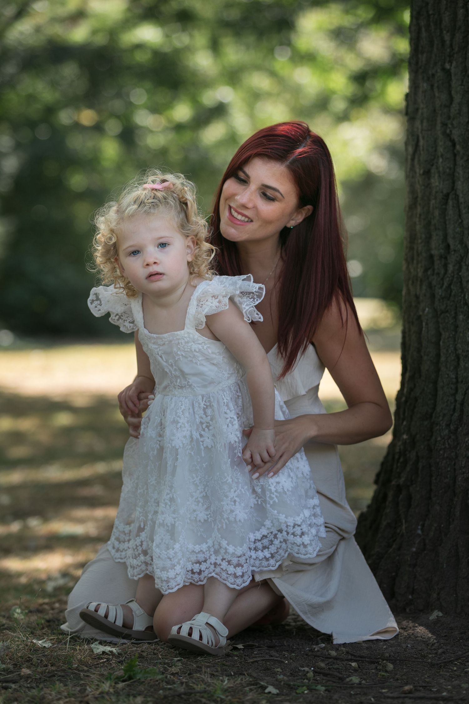 A smiling red-haired woman crouches beside a curly-haired toddler girl, both wearing white dresses in a sunny park.