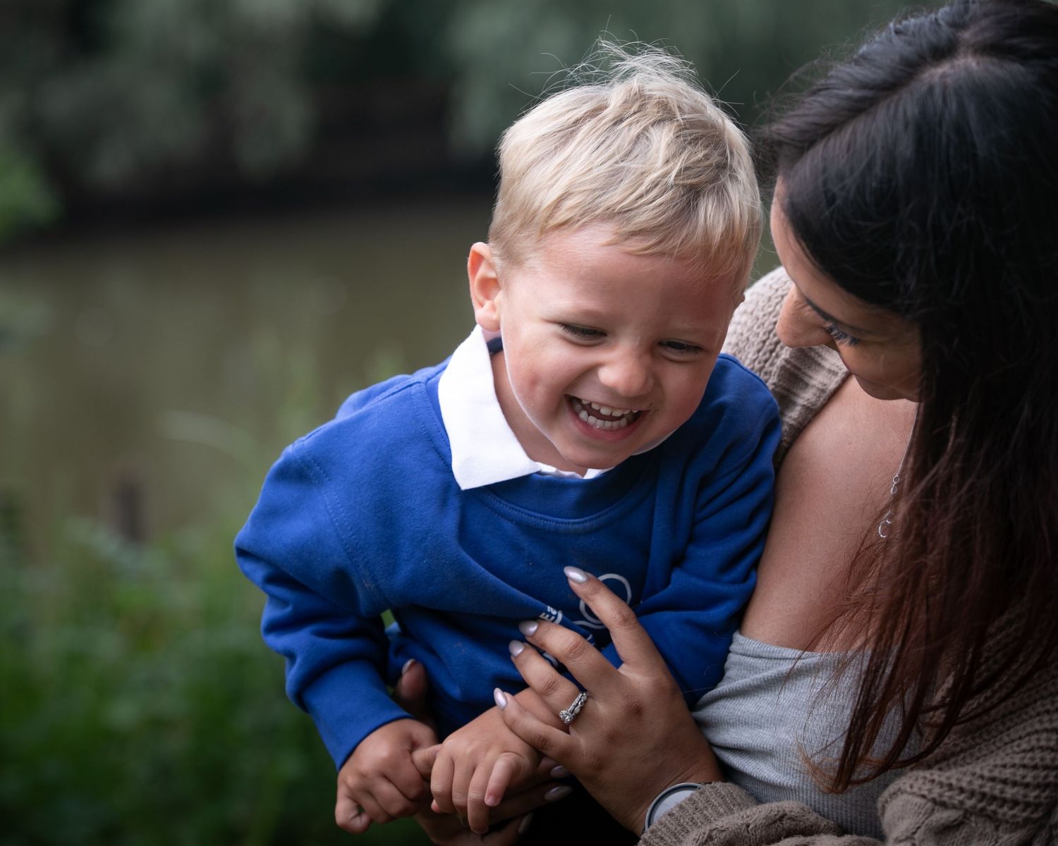 A joyful toddler in a blue sweater laughs while being held by a dark-haired woman outdoors near greenery.