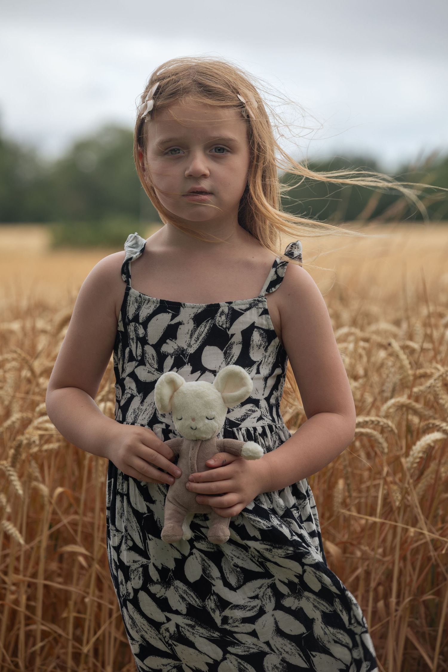 Young girl in floral dress holding stuffed mouse toy standing in golden wheat field on cloudy day.