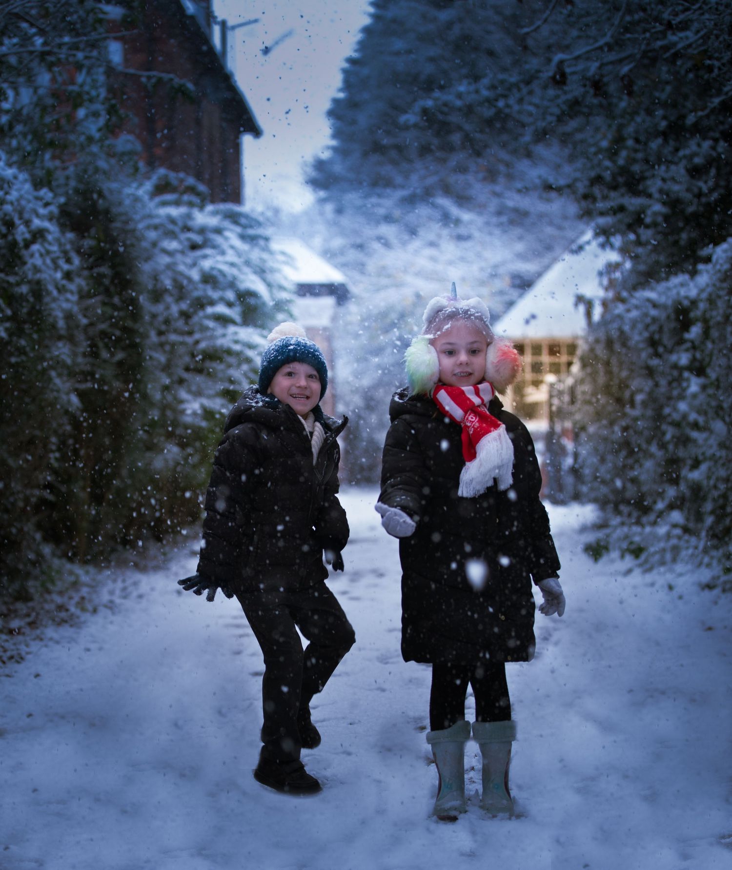 Two children play in falling snow on a winter street lined with snowy trees.