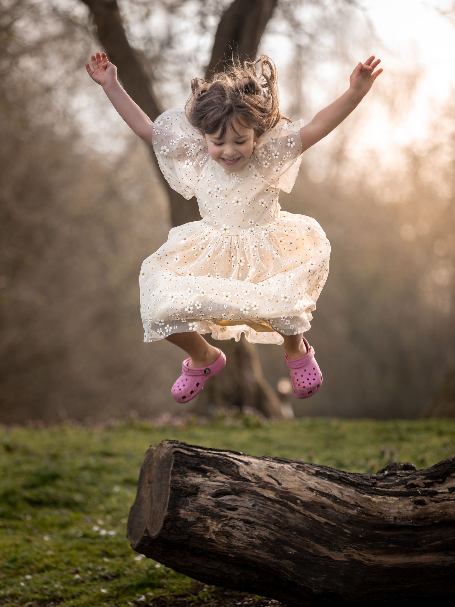 A joyful young girl in a white dress and pink Crocs leaps off a log in a sunlit woodland setting. east park Hull