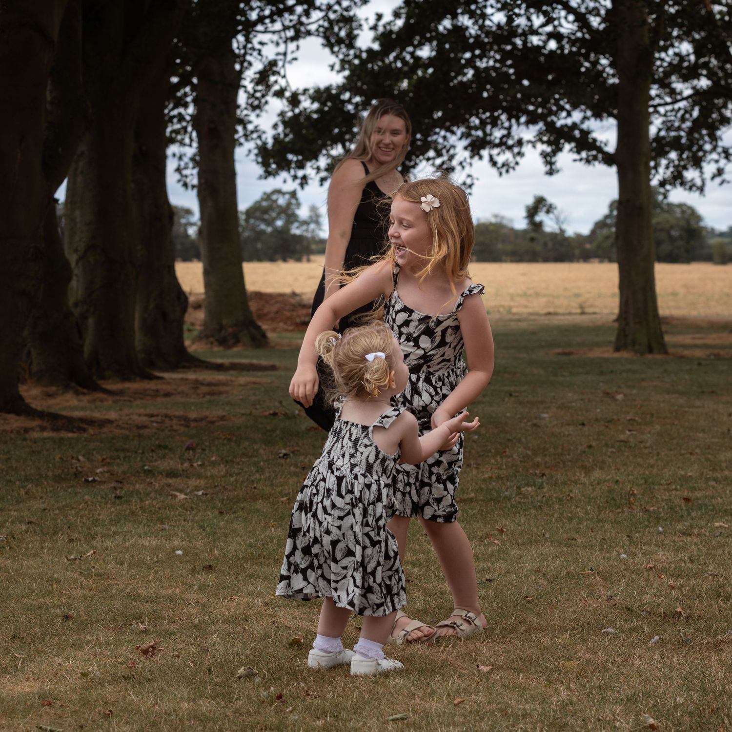 Two girls in matching black and white floral dresses play outdoors near trees, with a woman smiling behind them.