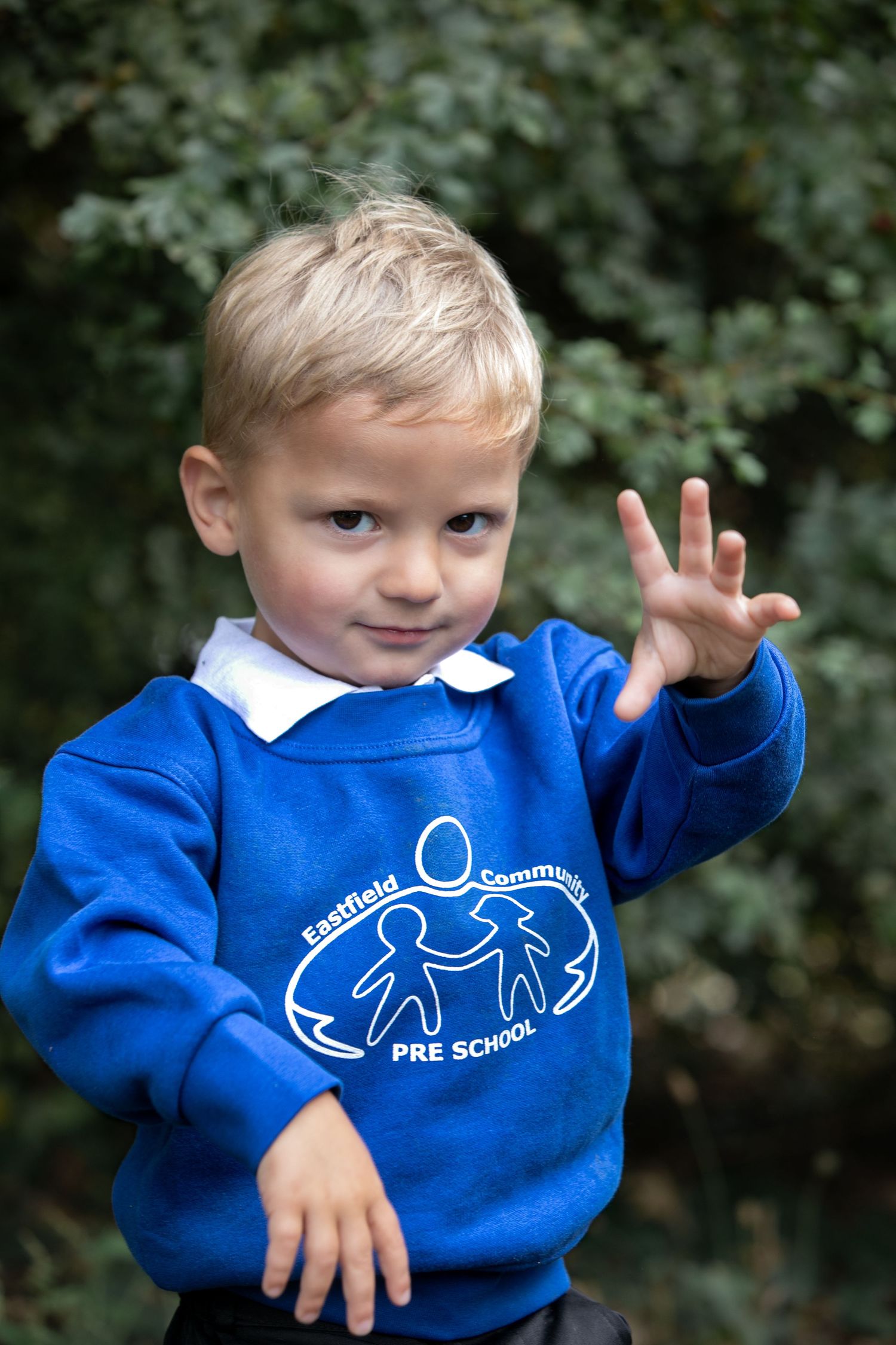 Young boy in Eastfield Community Pre School blue sweatshirt holding up three fingers outdoors.