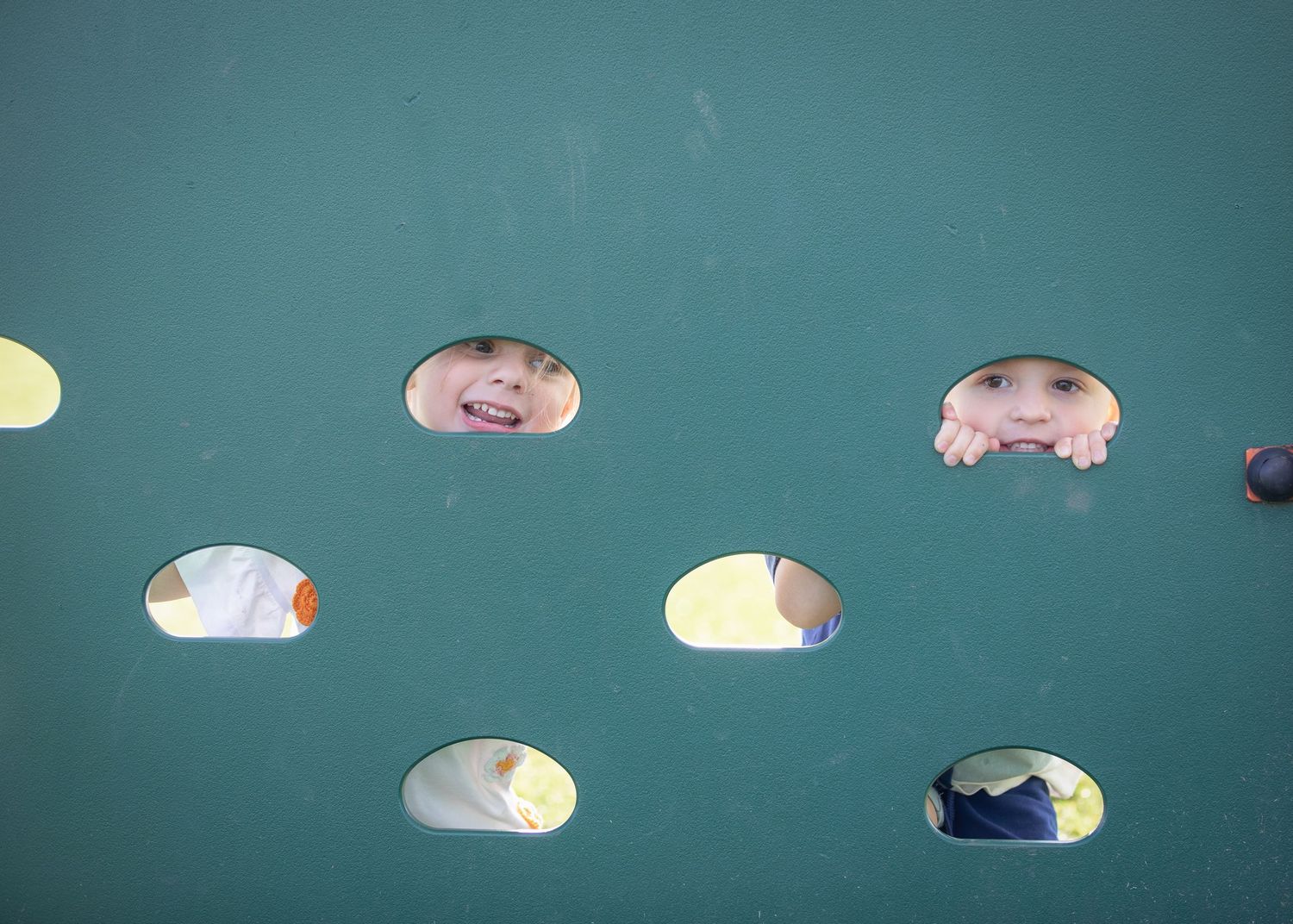 Children peeking their faces through oval holes in a teal playground wall, smiling and playing.