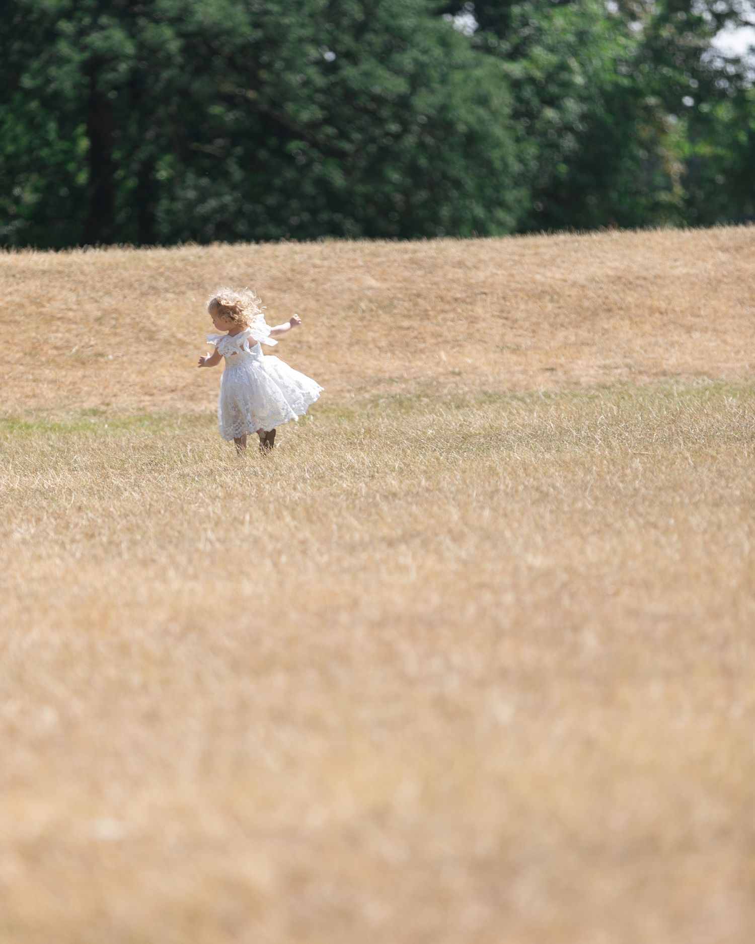A young girl in a white dress and hat twirls joyfully in a golden summer meadow with trees in the background.