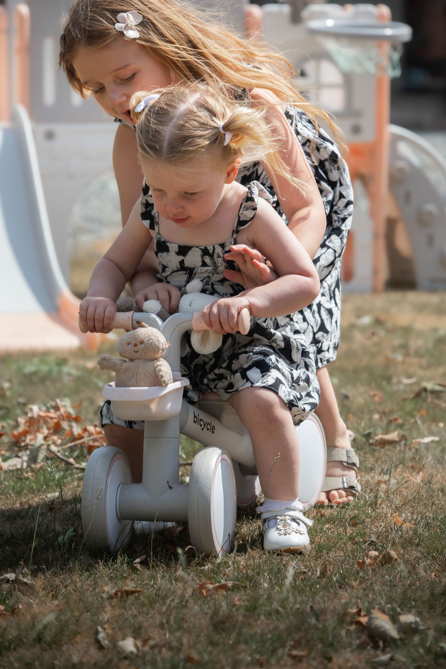 Two young girls playing outdoors with a white toy ride-on bike, one helping the other steer in a sunny park.