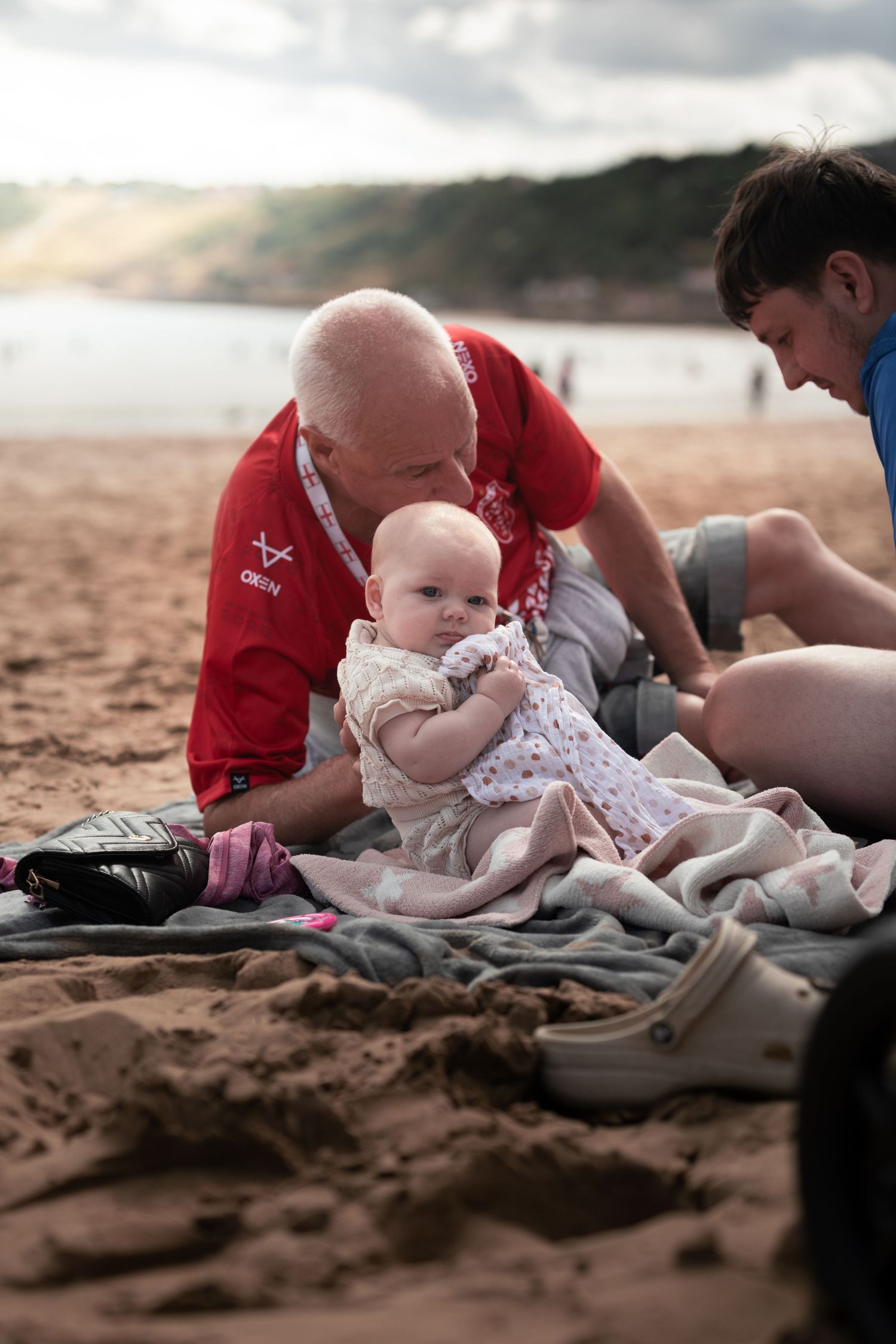 An older man in a red shirt and a teenager tend to a baby on a blanket on a sandy beach.

Scarborough