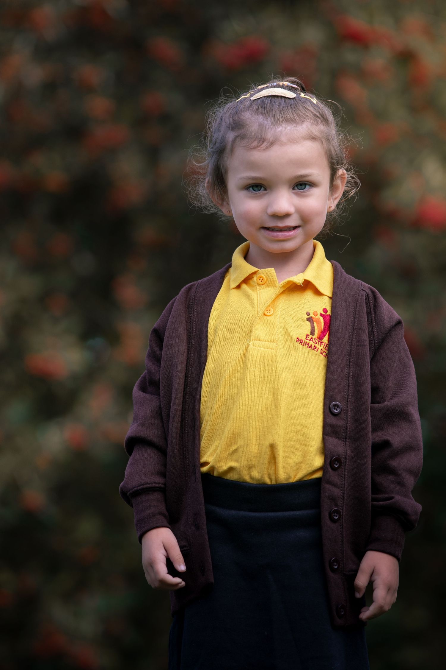 Young girl in yellow school polo shirt and brown cardigan smiling outdoors with red berries in background.