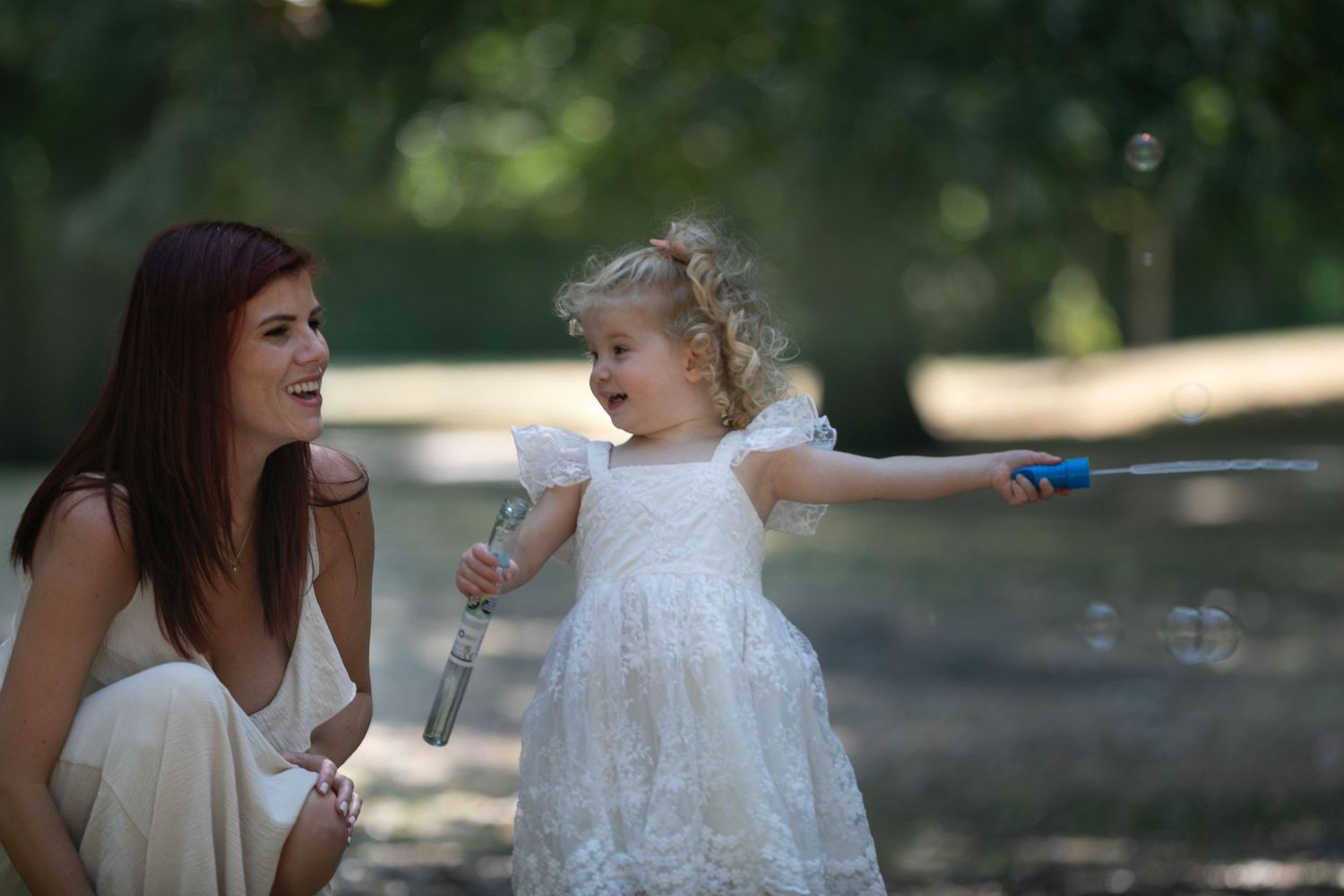 A woman in white dress laughs as a curly-haired toddler girl blows bubbles with a wand in a sunny park.