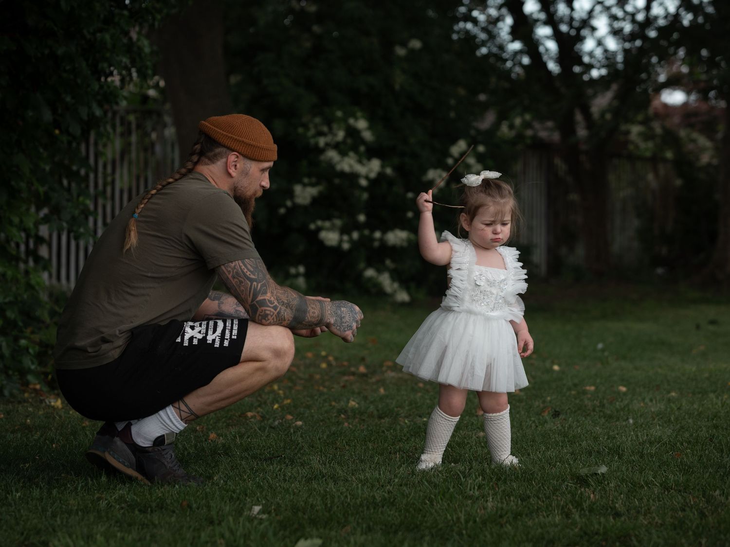 Tattooed father crouching to interact with toddler girl in white tutu dress on green lawn at dusk.

East Park Hull