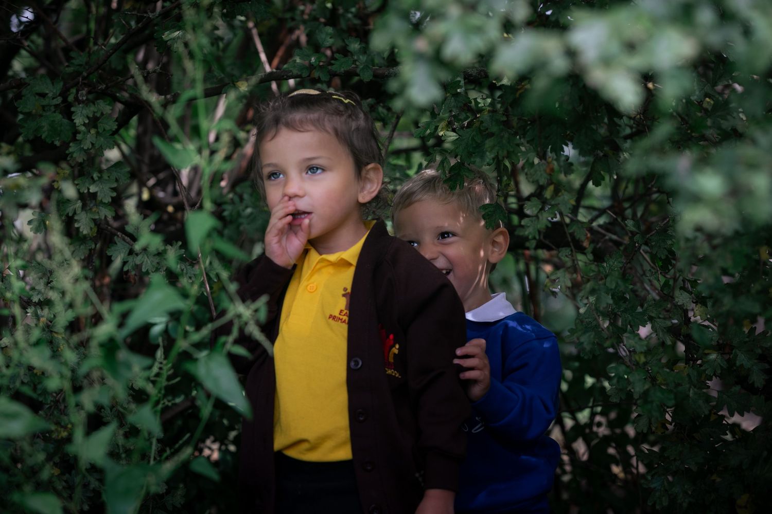 Two young children playing among green bushes and trees, one wearing a yellow shirt and one in a blue jacket.