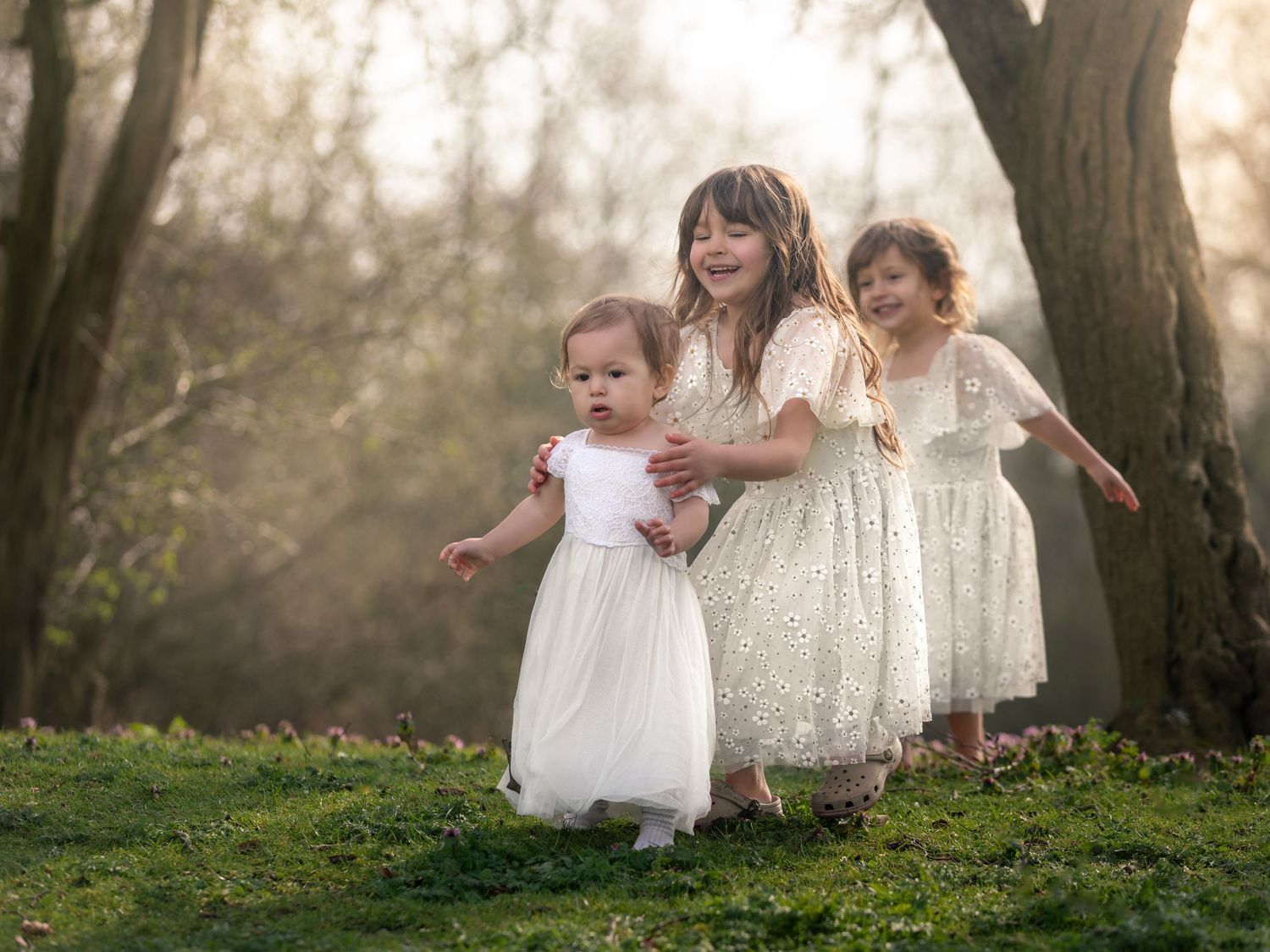 Three young girls in white lace dresses playing outdoors in a sunlit forest setting.