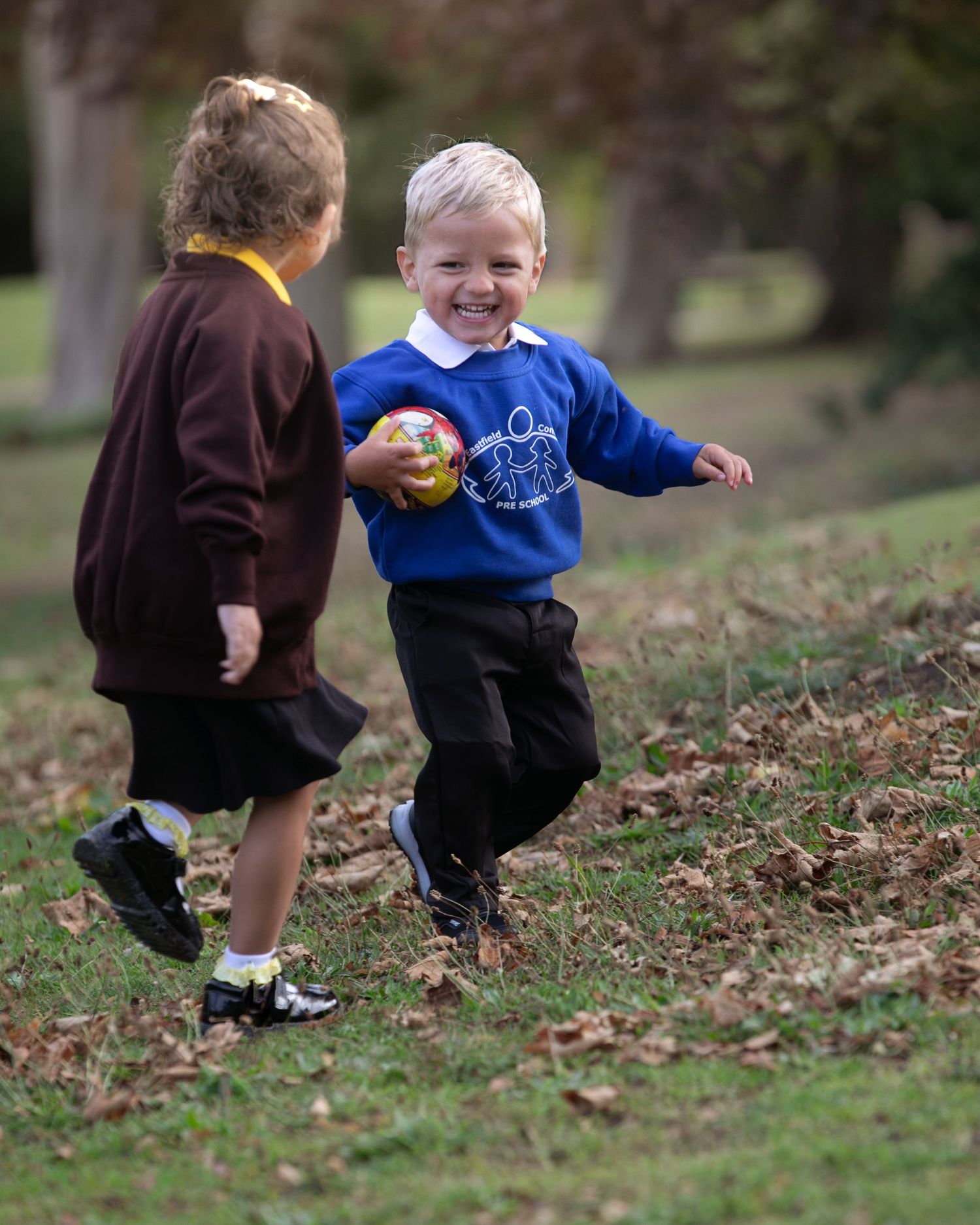 Two young children in school uniforms playing outdoors, with a smiling boy holding a ball running on grass.