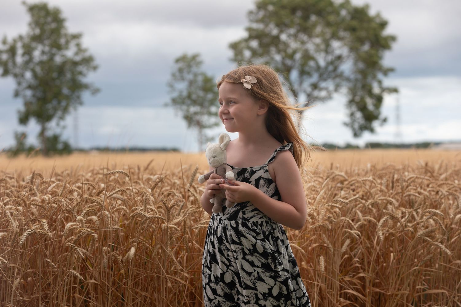 Young girl in floral dress holding wheat stalk in golden grain field on cloudy day.