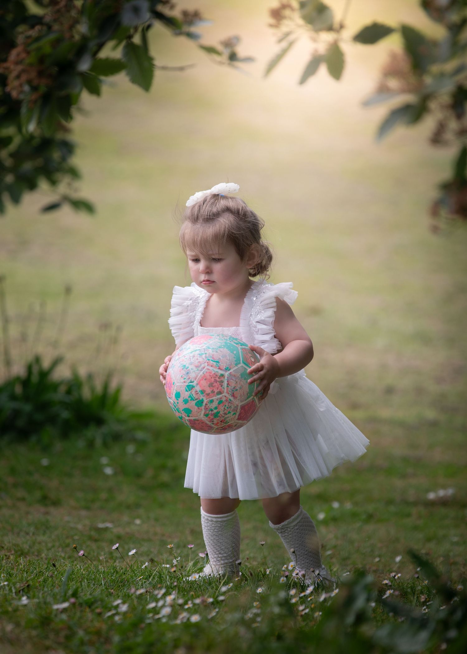 Toddler girl in white tutu dress holding a pink and green soccer ball outdoors in a garden setting.
East Park Hull