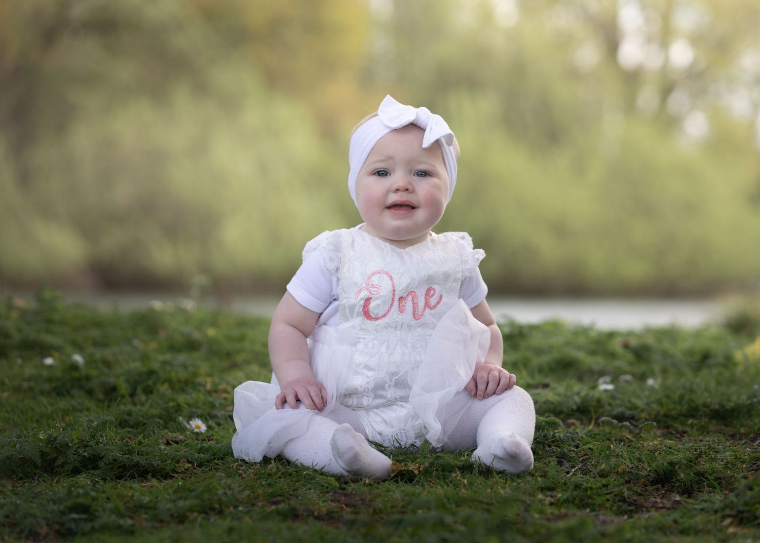 A baby girl in a white 'One' romper and bow headband sits on green grass outdoors for her first birthday photo.