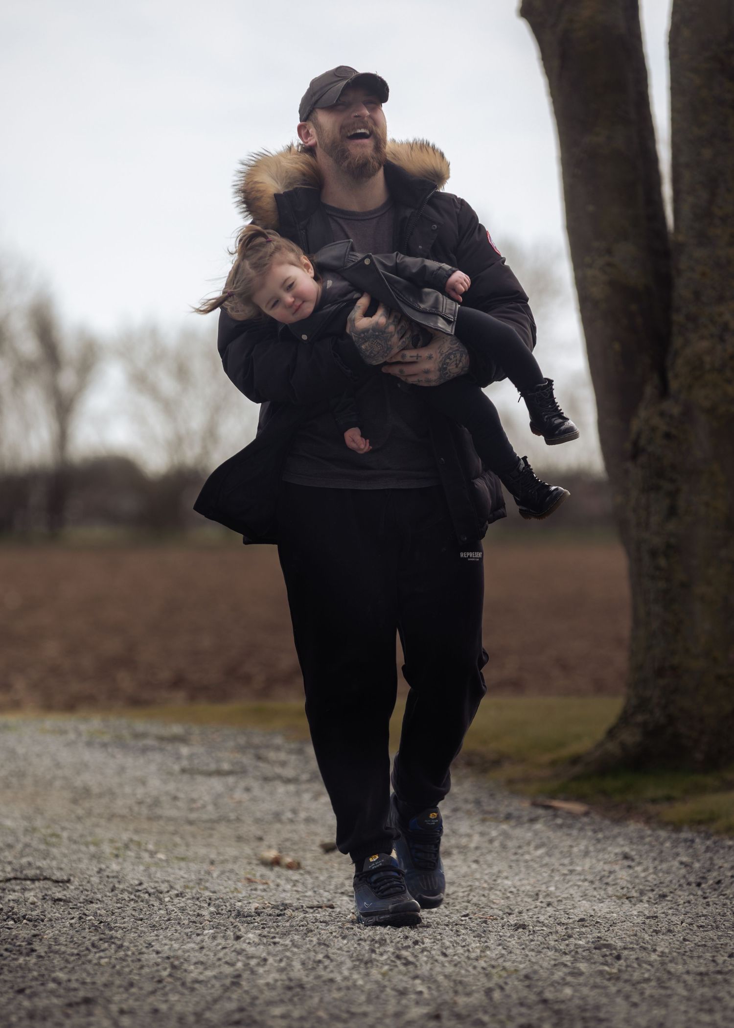 A smiling bearded man carries two young children while walking along a gravel path in a park on a cloudy day.