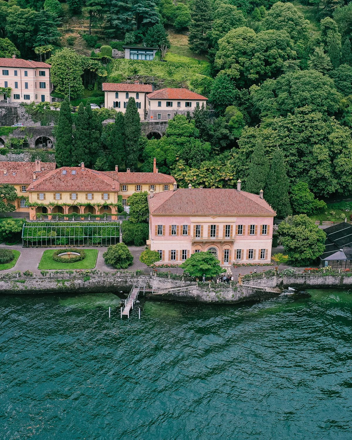 Aerial view of elegant Italian lakeside villa with manicured gardens surrounded by lush green hillside trees.