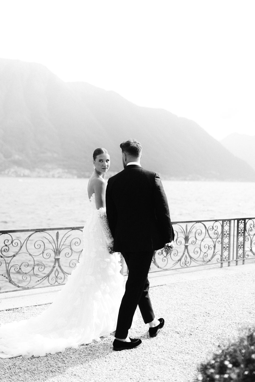Bride and groom walking along ornate iron railing overlooking a lake with mountain backdrop in black and white.