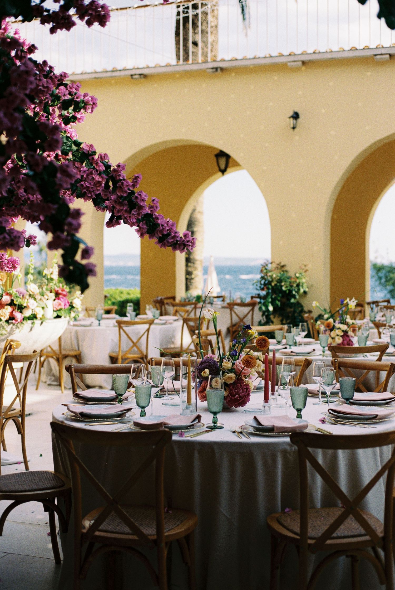 Outdoor wedding reception at a yellow villa with purple bougainvillea, round tables, cross-back chairs, and ocean views.