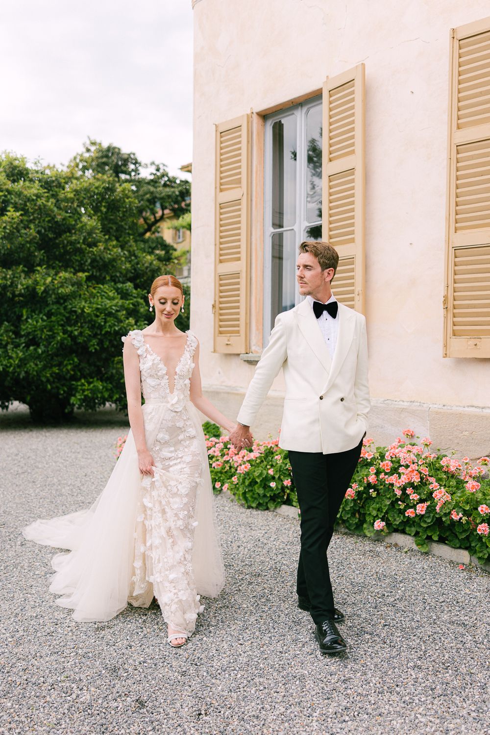 Bride in lace gown and groom in white tuxedo hold hands walking outside an Italian villa.