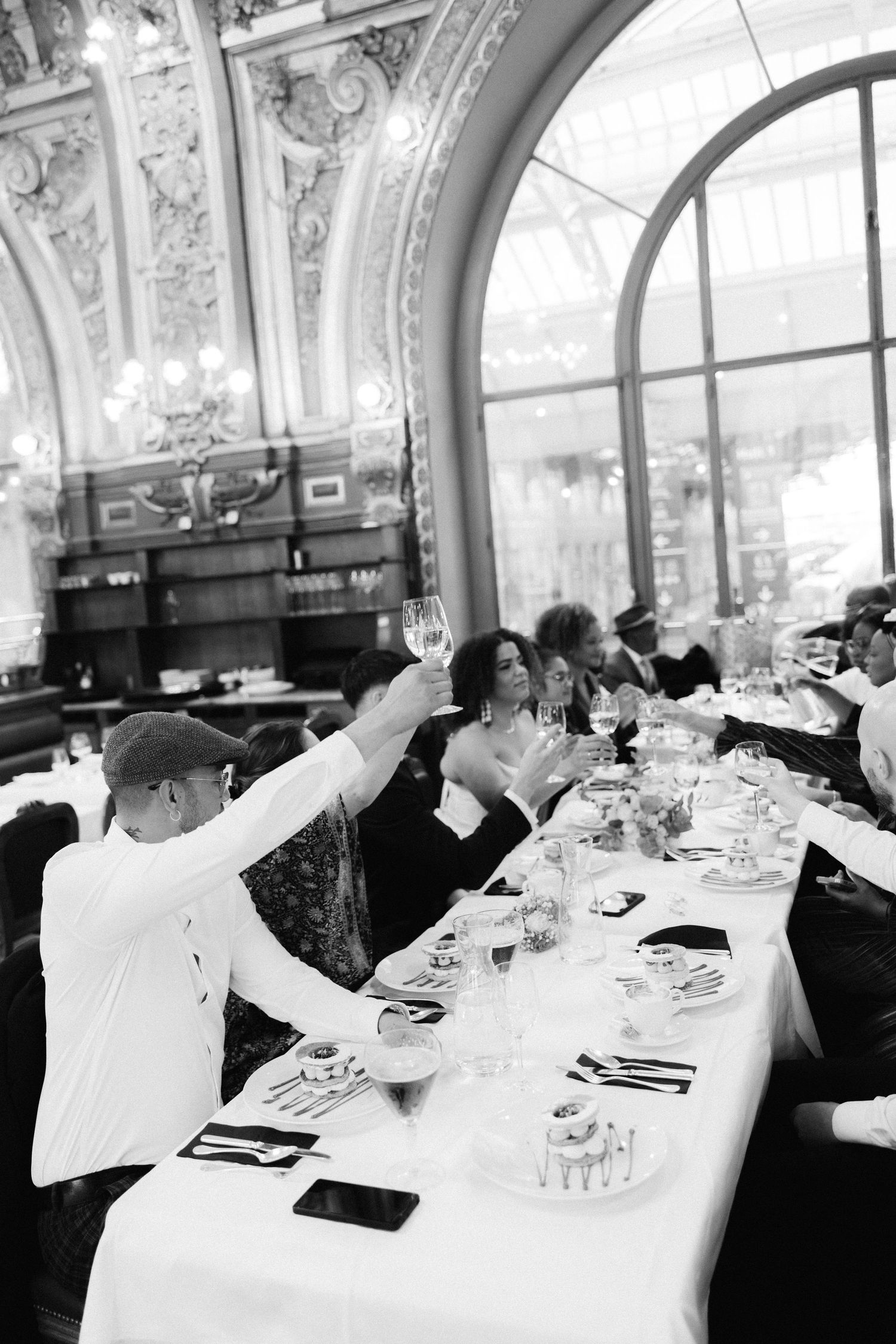 Black and white photo of wedding guests raising champagne glasses in toast at ornate Parisian restaurant reception.