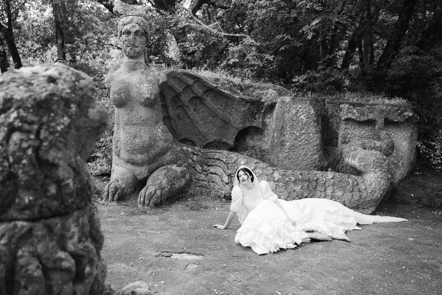 Bride in white wedding gown posing beside ancient stone sculptures in a wooded garden setting.