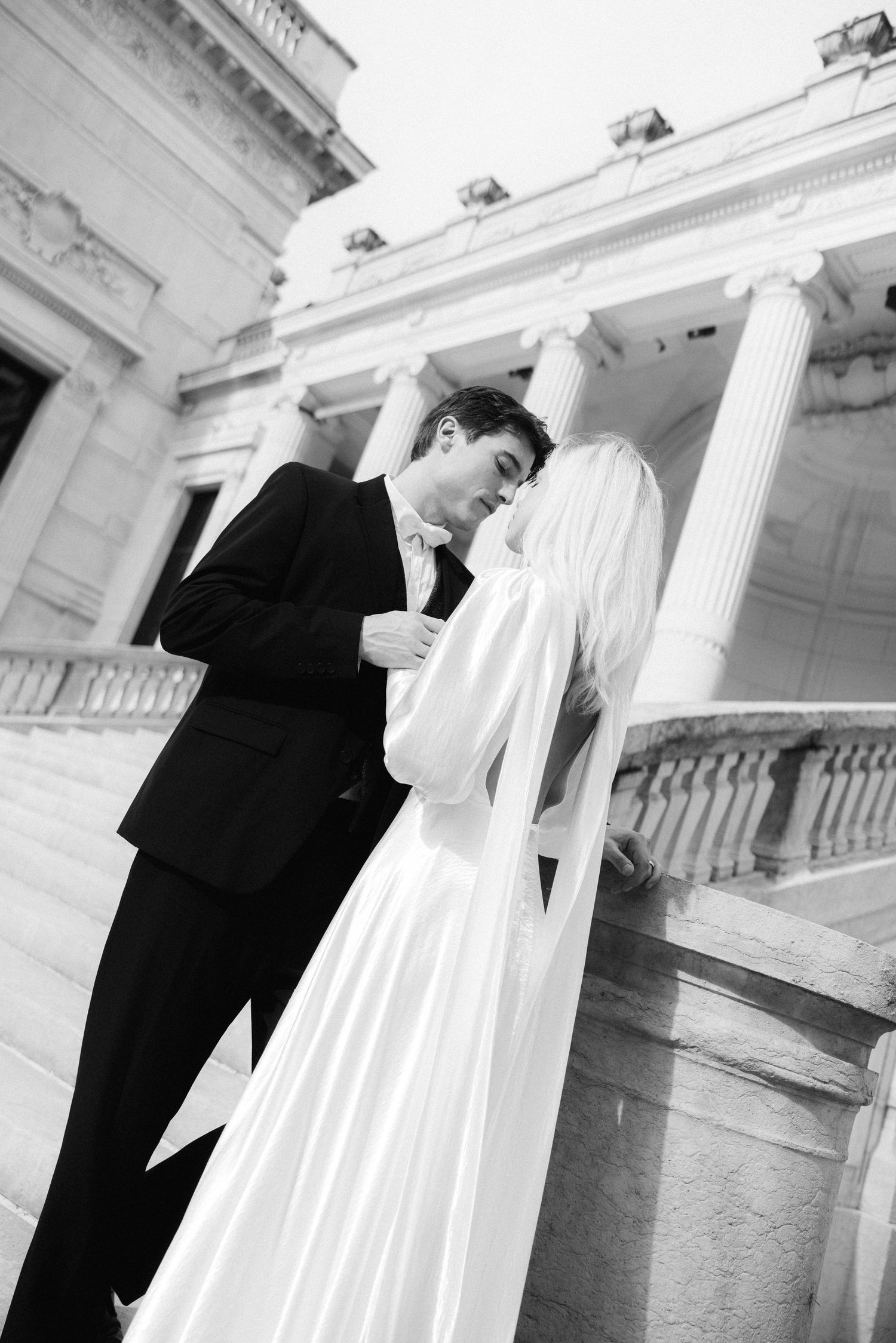 Bride and groom share a kiss on neoclassical building steps, bride wearing flowing white satin gown.
