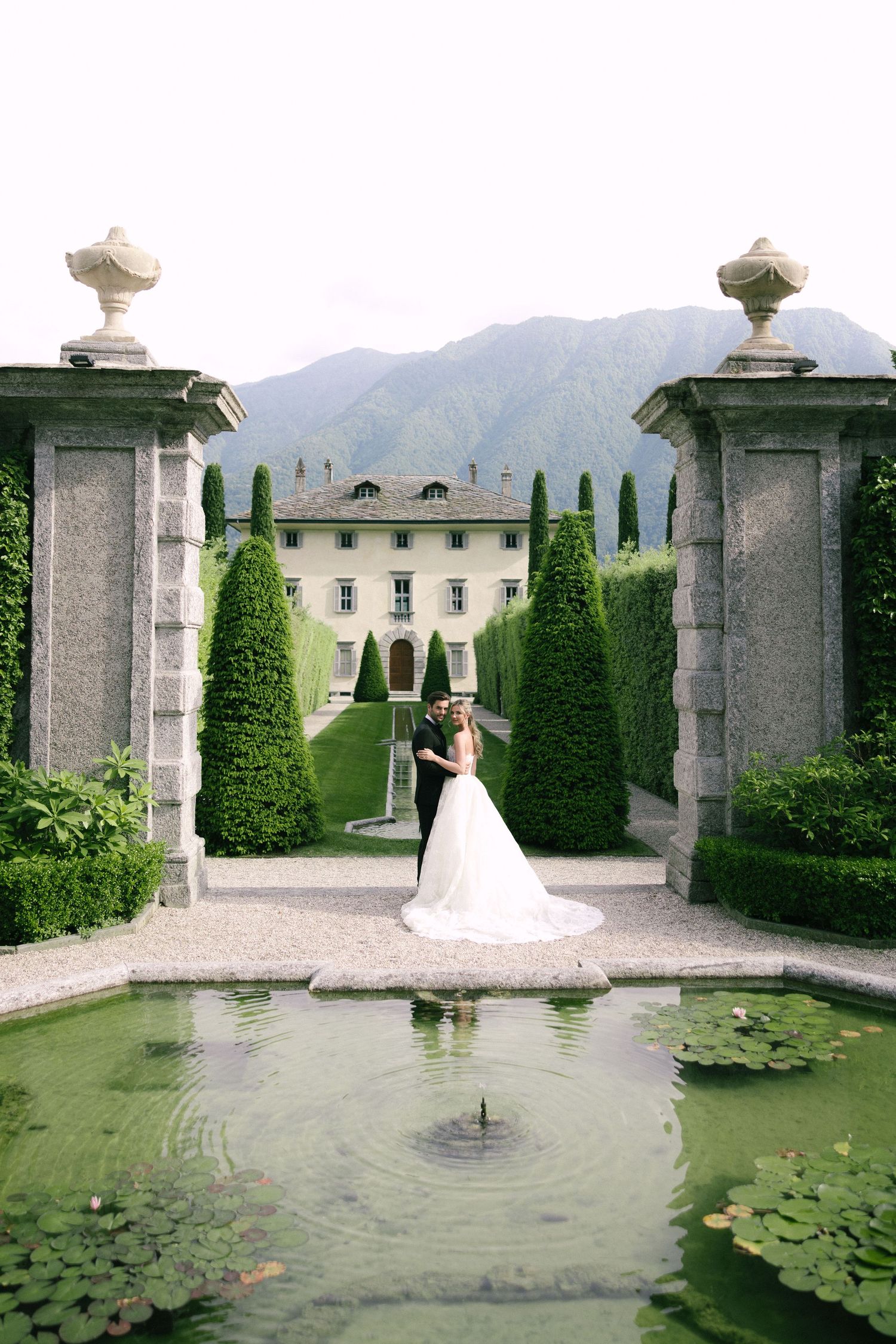 Bride and groom embracing in formal Italian villa garden with lily pond, topiary trees, and mountain views.