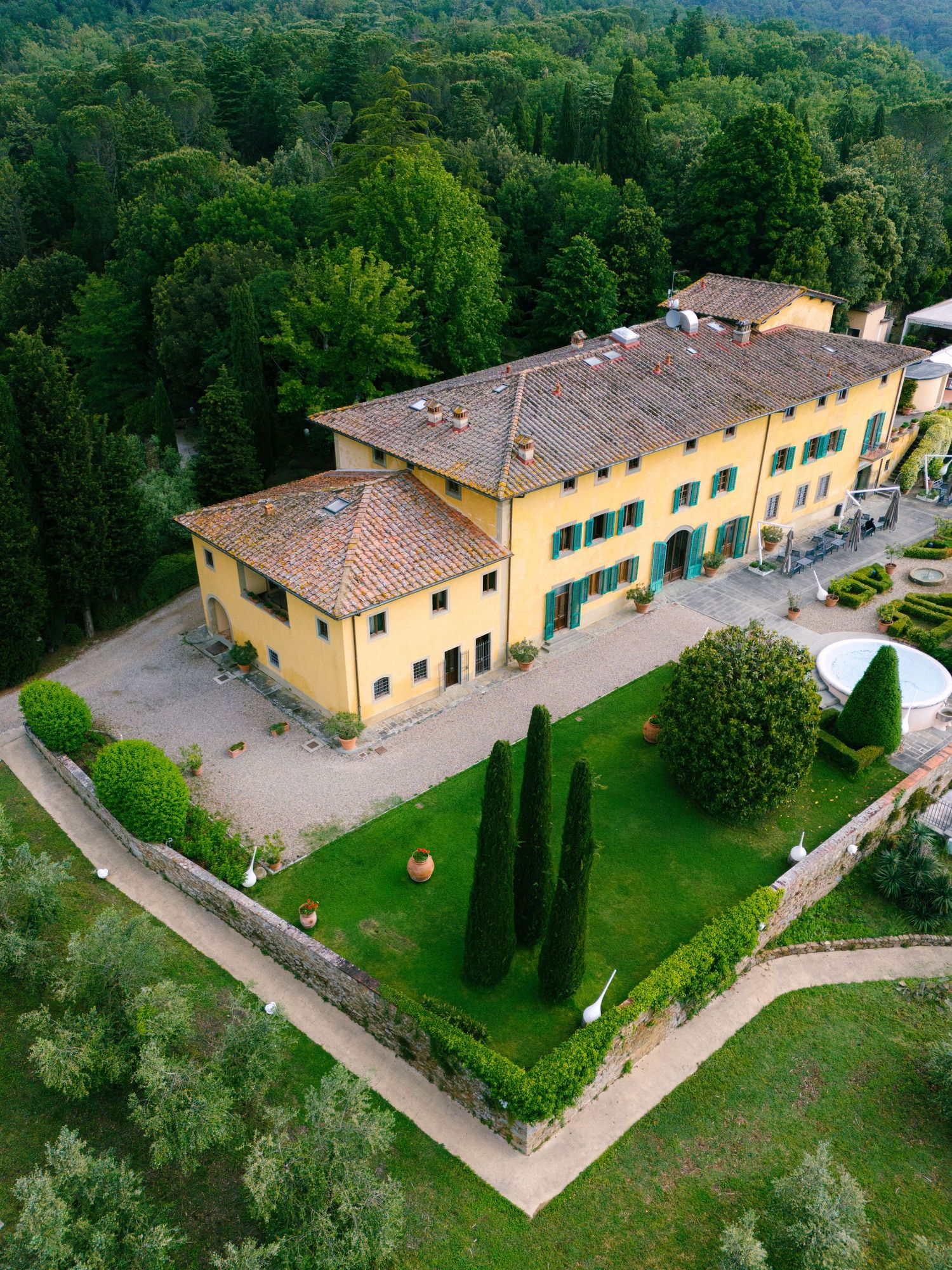 Aerial view of elegant yellow Tuscan villa surrounded by cypress trees, manicured gardens, and lush forest.