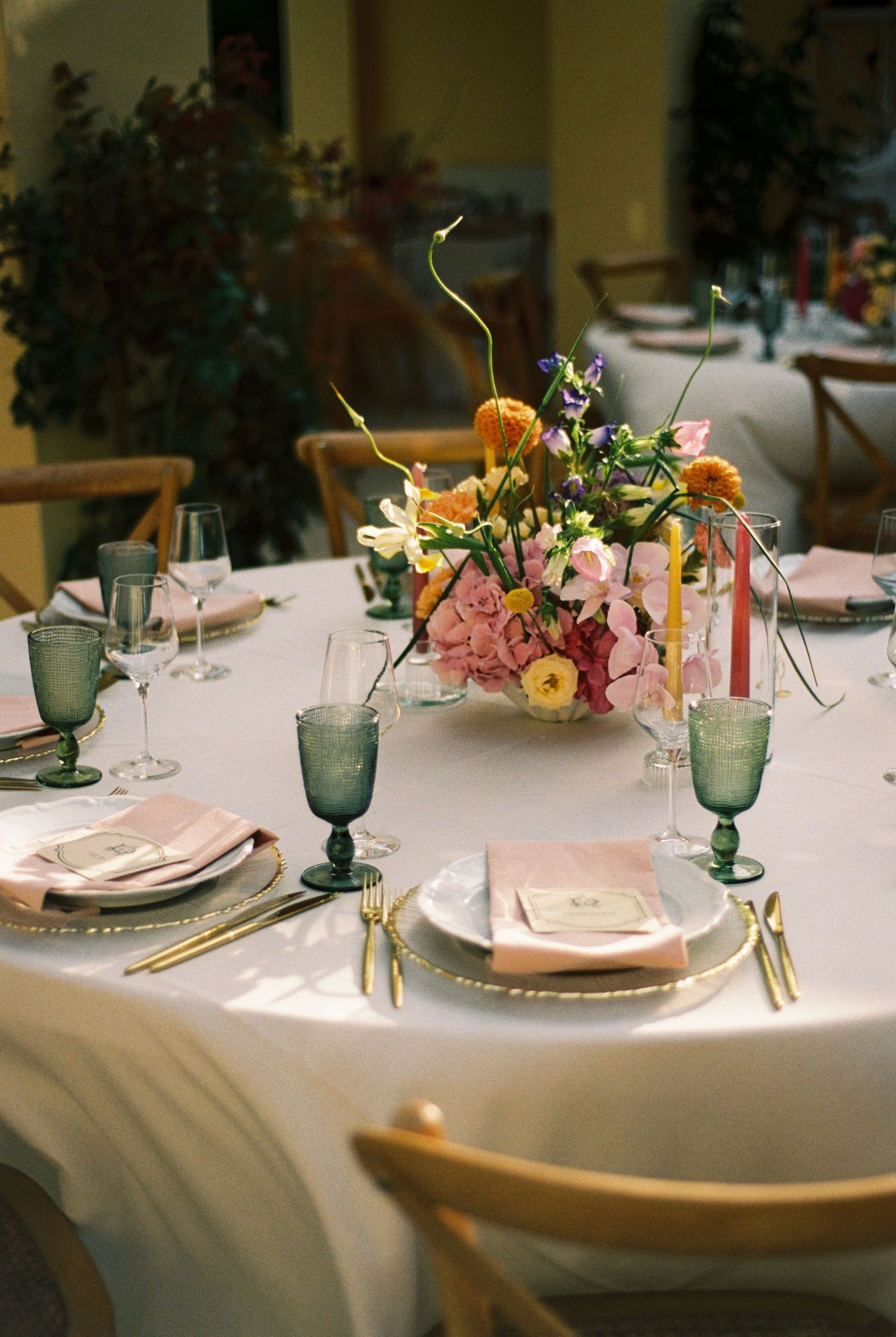 Colorful wedding table setting with vibrant floral centerpiece, green goblets, gold cutlery, and pink napkins indoors.