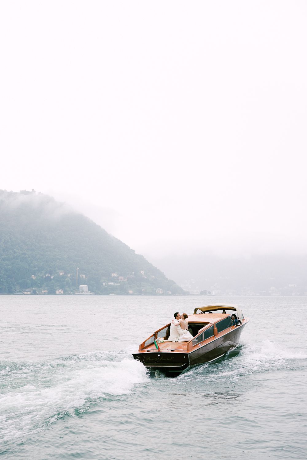 Bride and groom riding a wooden speedboat on misty Lake Como, Italy.