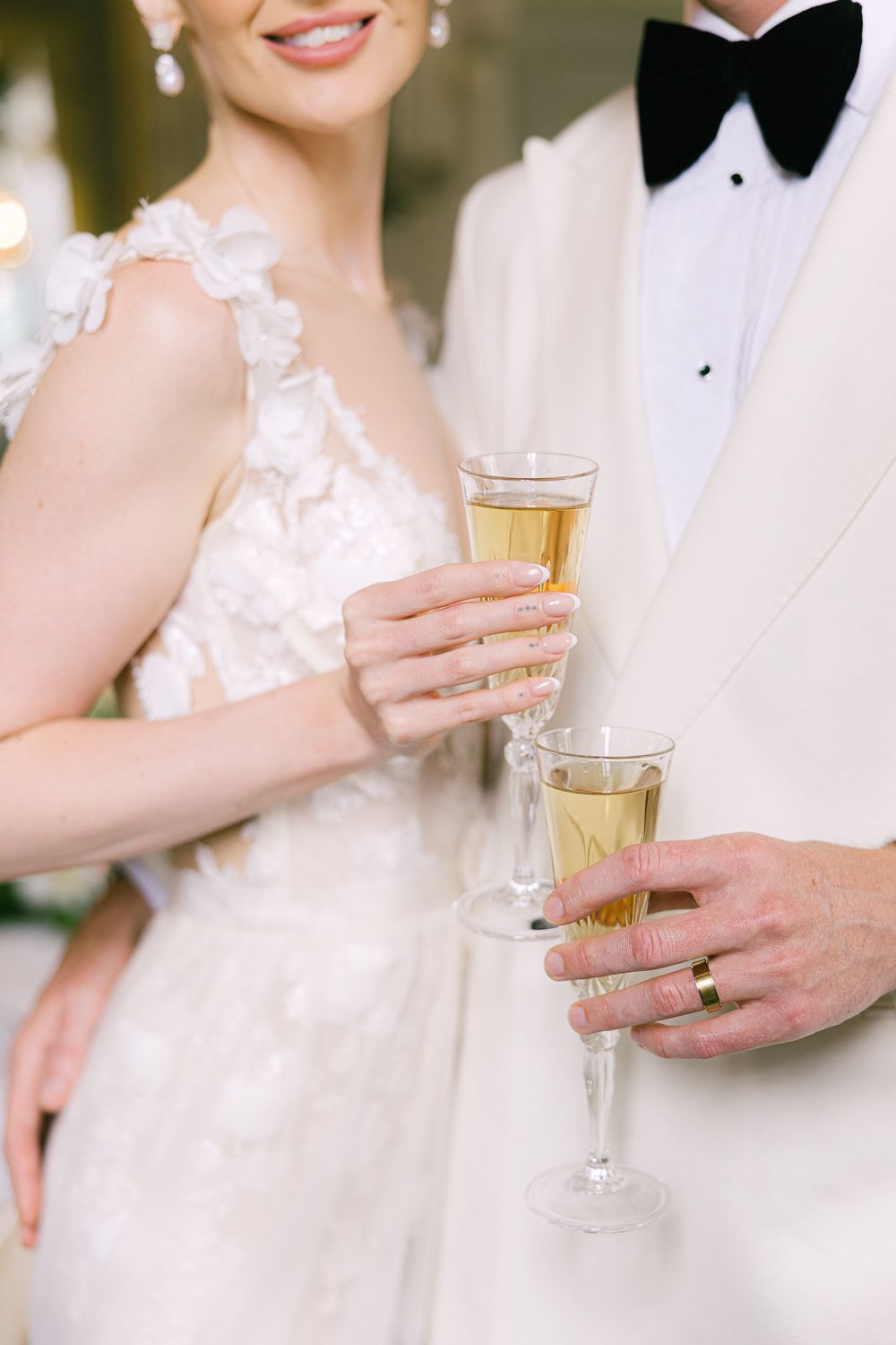 Bride and groom in white tuxedo holding champagne flutes with gold wedding rings visible at elegant reception.